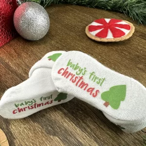 Flat lay of white baby socks that say “Baby’s First Christmas” in red and green with a small Christmas tree graphic, surrounded by Christmas decorations on a wooden background