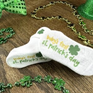 Baby's first st. Patrick's Day socks with green shamrock graphic laying on wood grain background surrounded by st. patrick's day decor (glittery green top hat, green bow tie, green shamrock necklaces)