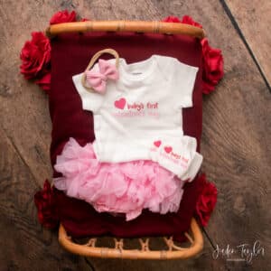 Flat lay of outfit set including bodysuit and matching socks with phrase "baby's first valentine's day" written in red and pink with a red heart graphic. Clothing set also includes pink ruffle bloomers and a pink hairbow on nylon headband. Outfit set is lying on a wooden mini bed draped with a red knitted blanket with a wood grain background with red roses peeking out around edges of bed. The bottom right corner has a logo with the words "Jaden Taylor photography"