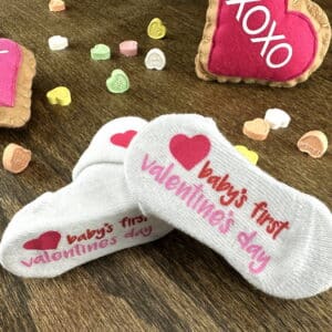 "Baby's First Valentine's Day" socks with red heart graphic all laying flat on wood grain background surrounded by conversation hearts candy and two larger felt versions of the conversation hearts candy
