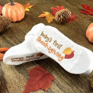 Babys First Thanksgiving socks with fall leaf graphic displayed on wood grain background amongst fall decor such as fall leaves, pine cones and pumpkins