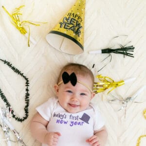 Baby's First New Years bodysuit with black top hat modeled by newborn girl with black hair bow and new year party favors