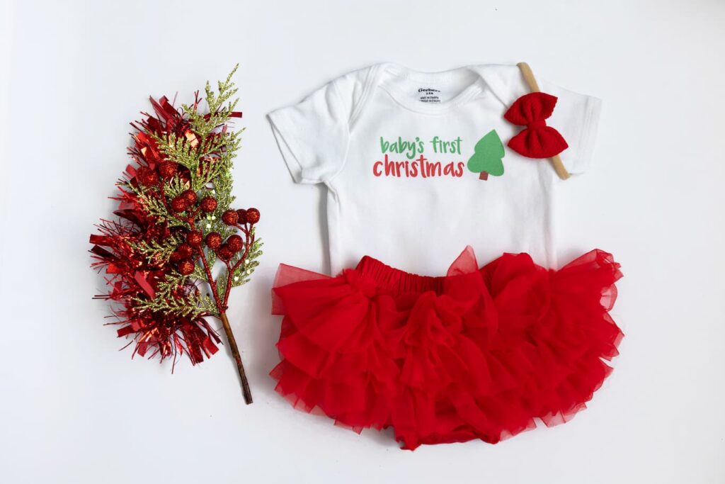 Flatlay of onesie with the phrase "baby's first Christmas" and Christmas tree graphic on bodysuit, with red hair bow and red ruffle bloomer all displayed next to festive Christmas greenery on white background