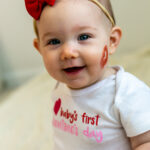 Smiling baby wearing "baby's first valentine's day" bodysuit and red hairbow with red lipstick kiss on baby's cheek, and cream colored background