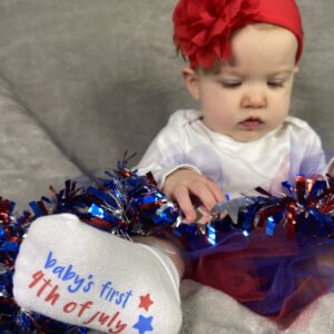 "Baby's First Fourth of July" socks modeled on older baby wearing white long-sleeved bodysuit, red flower headband, sitting up and looking at red, silver and blue garland, all on grey background