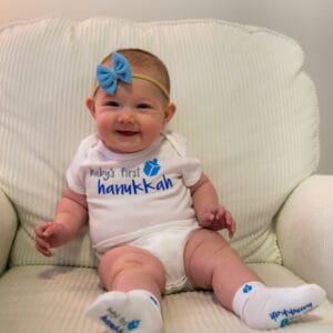 Baby's First Hanukkah socks and bodysuit with blue dreidal graphic and hairbow on smiling baby sitting on cream striped armchair