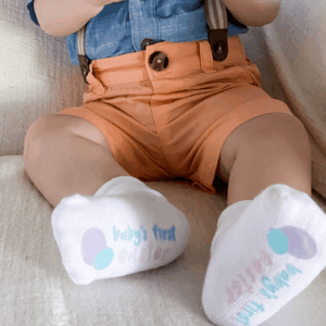 Close up picture of boy wearing "Baby's First Easter" socks, orange pants and a blue button up shirt, sitting on cream textured background