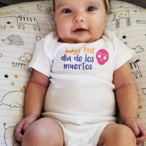 Baby's First Dia de los Muertos bodysuit and sock set with sugar skull graphic, modeled on baby laying down on animal boppy wearing orange hairbow