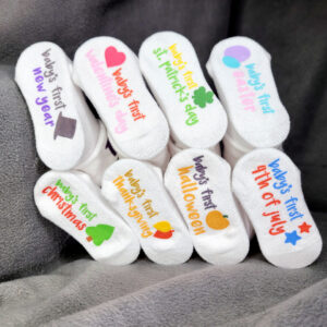 8 major American holiday "Baby's First" sock set with socks upside down with toes facing inwards towards each other to form a circle. The following text shows on the bottoms of the socks: Baby's first New Year, Baby's First Valentine's Day, Baby's First St. Patrick's Day, Baby's First Easter, Baby's First Christmas, Baby's First Thanksgiving, Baby's First Halloween, Baby's First 4th of July.