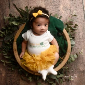 Newborn baby with open eyes wearing baby's first st patrick's day bodysuit and socks with yellow hairbow and yellow ruffle bloomers, laying in wooden basket and green fabric on top of round green leaves and wood grain backdrop. Bottom corner has the logo for Jaden Taylor Photography