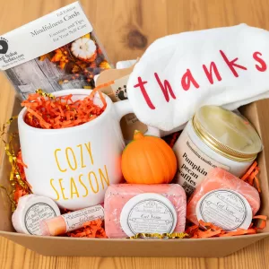 fall-themed self-care spa basket displayed in a cardboard tray on a wooden background, featuring a candle, bath bomb, mindfulness cards, cozy socks, mug, stress ball, lip balm, soap, and scrub.