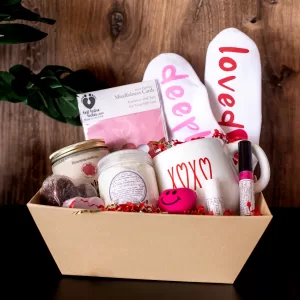 Love-themed self-care spa basket neatly arranged in a cardboard tray on a wooden background, featuring a heart-shaped bath bomb, salted caramel brown sugar soap, coconut vanilla shimmer lotion, cherry lemon lip balm, watermelon pineapple lip gloss, Bohemian Rose or Strawberry Cheesecake candle, love-themed mindfulness cards, heart stress ball, cozy socks, and a Sweet, Kisses, or Love mug.